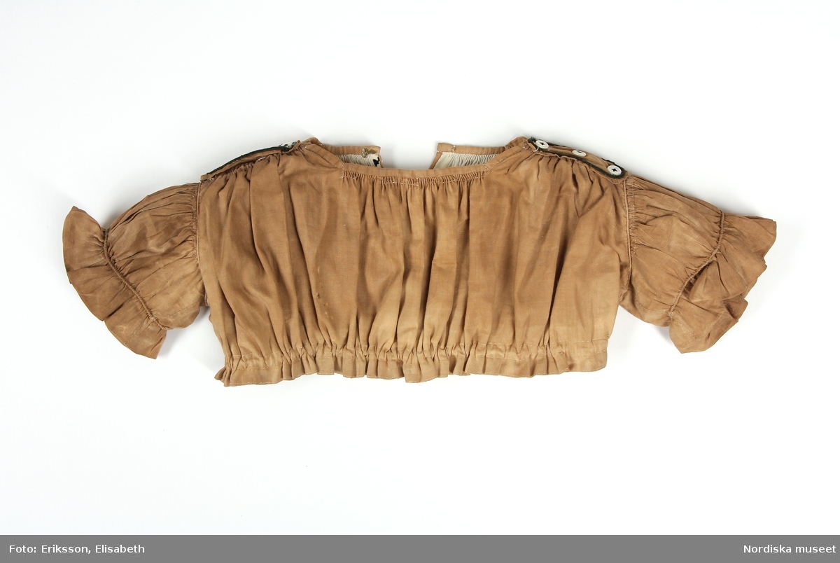 Huvudliggare:
"Barnklädning, bestående av: liv, kjol och skärp av gult bomullstyg med svarta och vita broderier. Början av 1860-talet:"