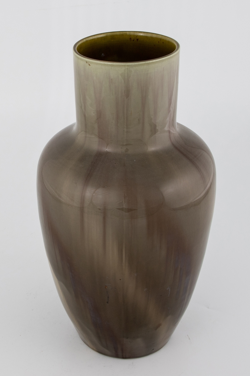 Vase i glasert flintgods. Urneformet koprus med sylindrisk hals. Vasen er dekket med grågrønn glasur, hvorpå rødbrune partier i form av rennende striper tidvis kommer til syne.