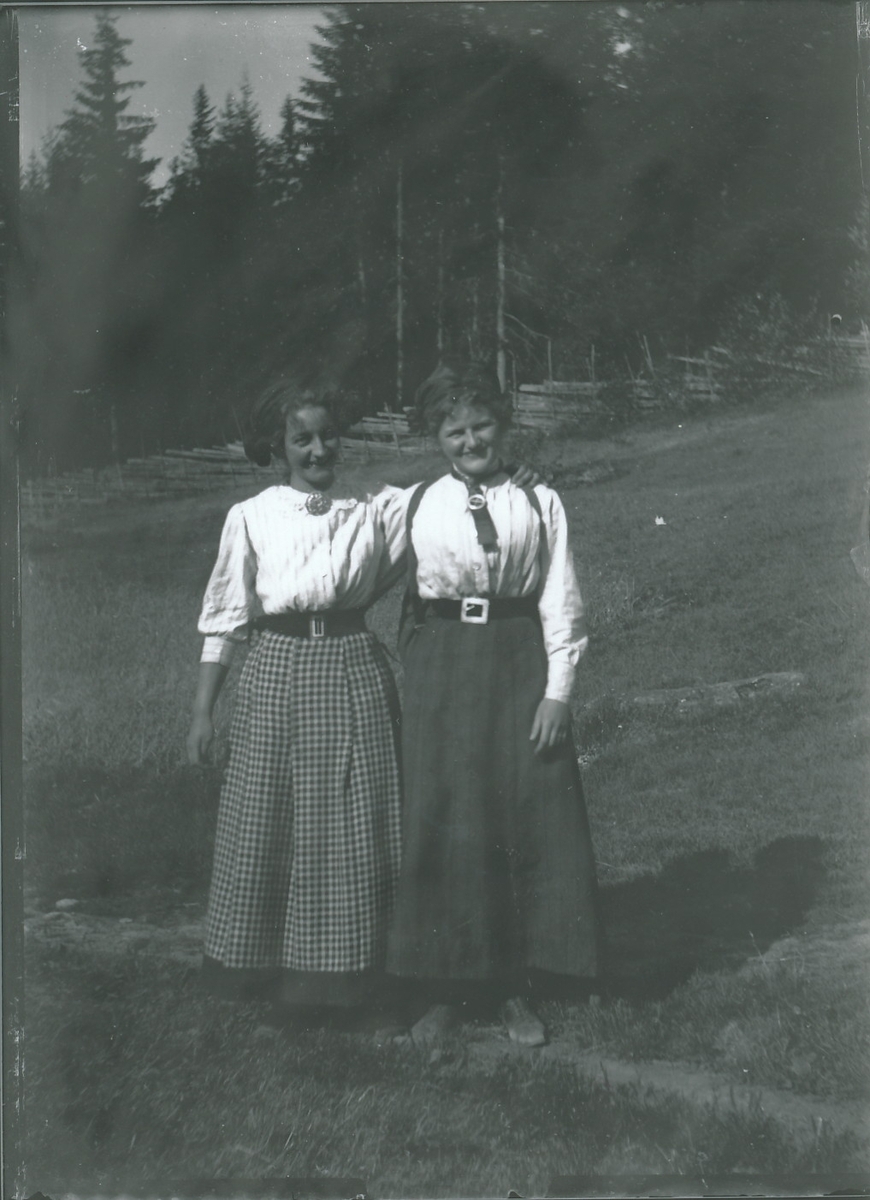 To unge kvinner arm i arm på grasbakke med skigard i bakgrunnen. Begge har hvite bluser med søljer i halsen, belte i livet, ankellange skjørt og hodeplagg.