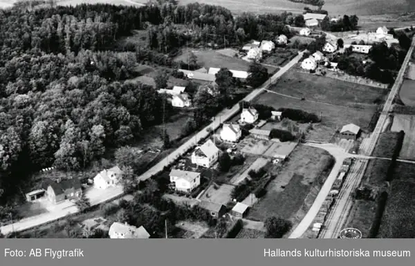 Vykort, "Flygfoto över Köinge". Enfamiljshus och byväg. Till höger ses en del av bangården till Falkenbergs Järnväg, "Pyttebanan". Lantgårdar med uthus. Odlingslandskap och skog.