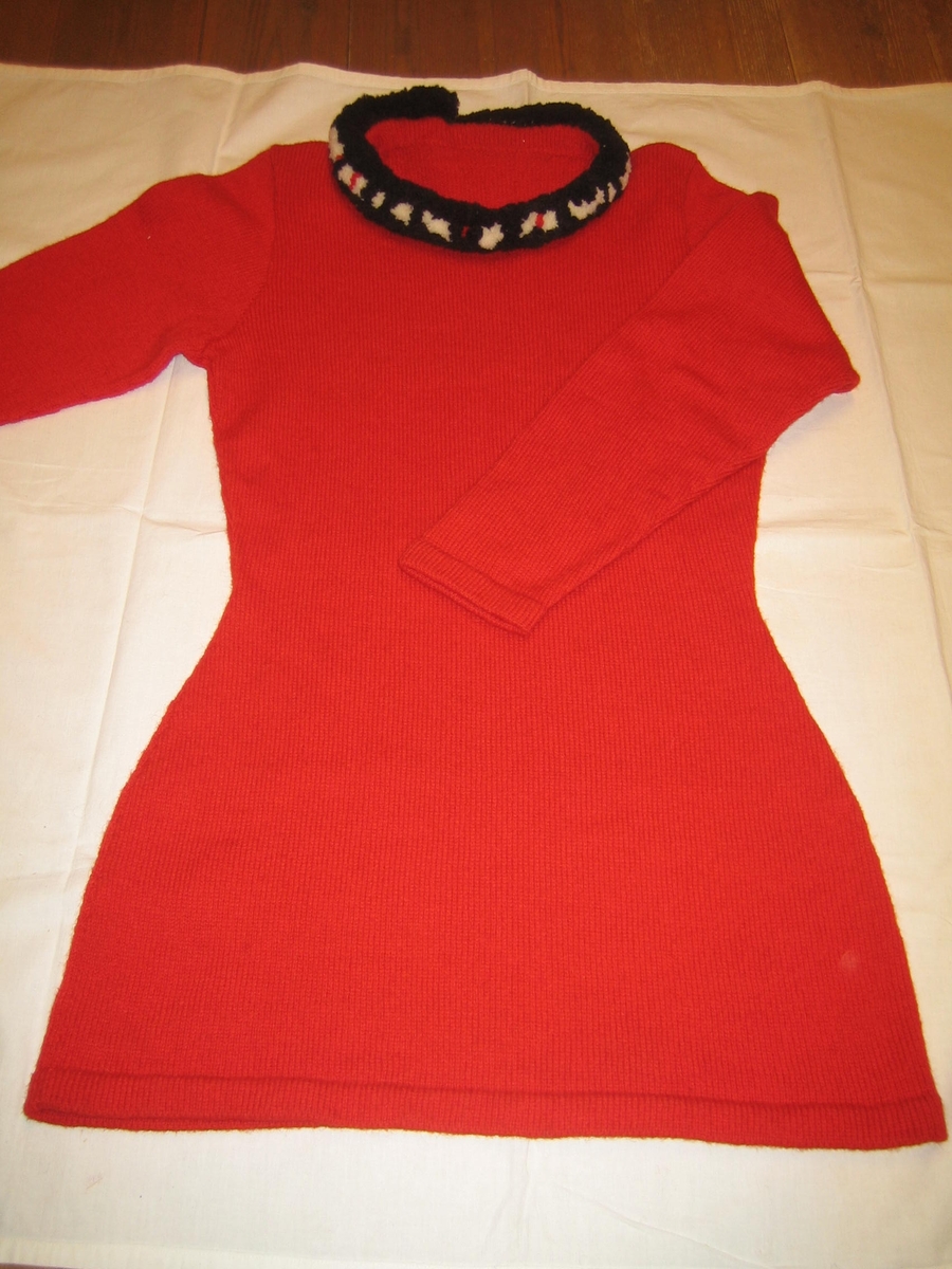 Röd slätstickad klänning med halskant som är rund med flossakrage i svart vitt och rött. Klänningsfåll och ärmavslut är i rätstickning dubbelvikt. Ärmen är isydd. På halskanten är fem bitar av kardborrband fastsydda.