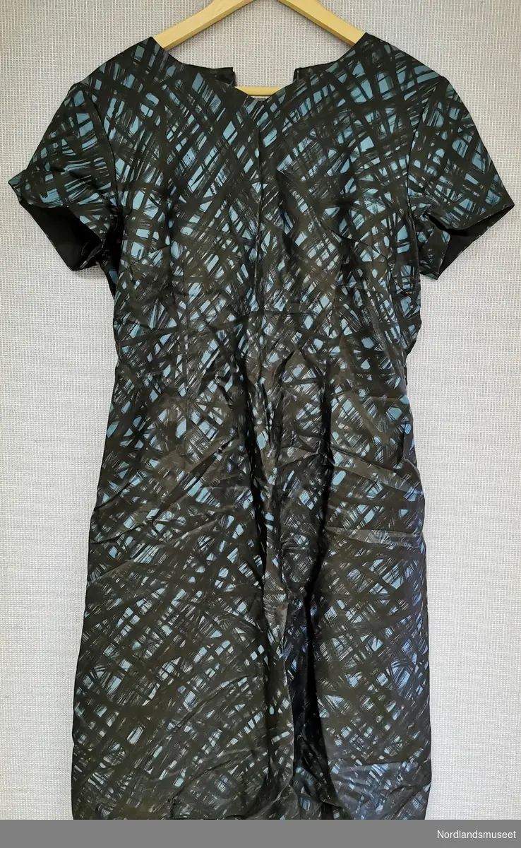 Petroliumsfarget kjole med svarte grafiske detaljer. 
Kjolen har glidelås i nakken og under venstre erme.
Den har et belte rundt livet.