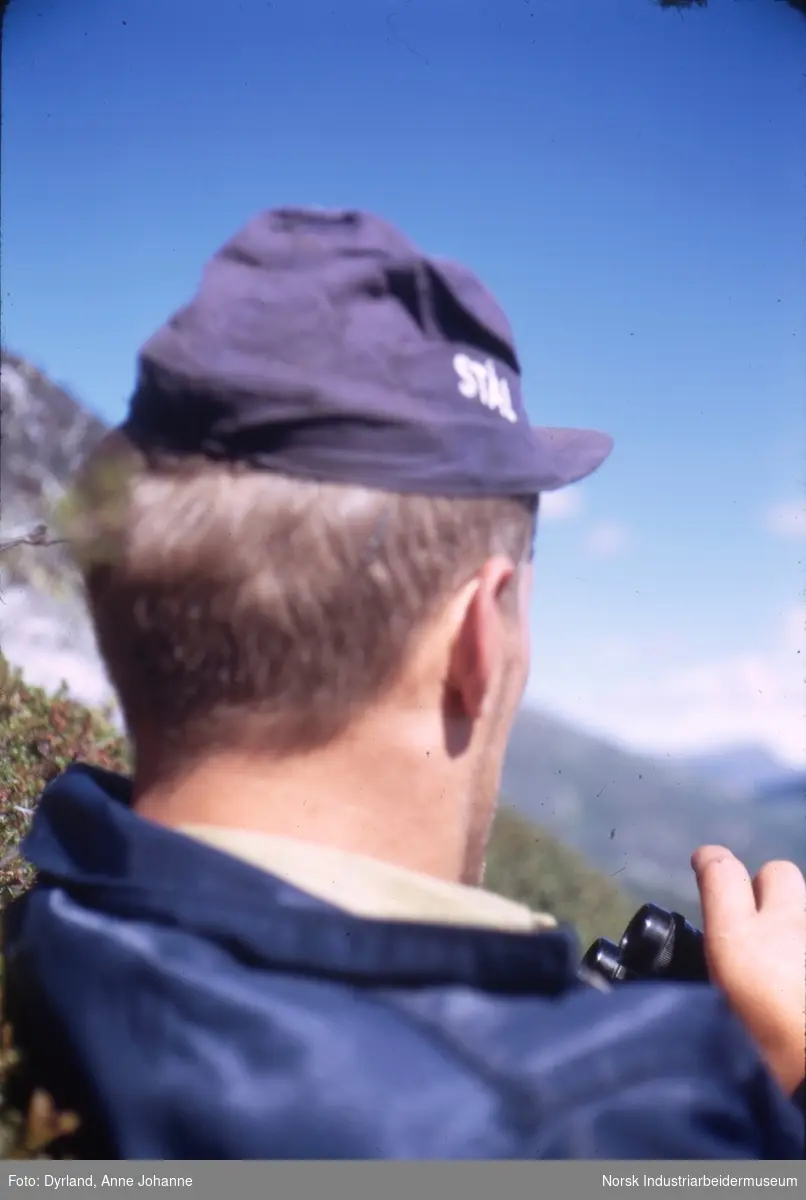 Bjørn Dyrland ligger i fjellet med kaps på hodet og kikkert på brystet.