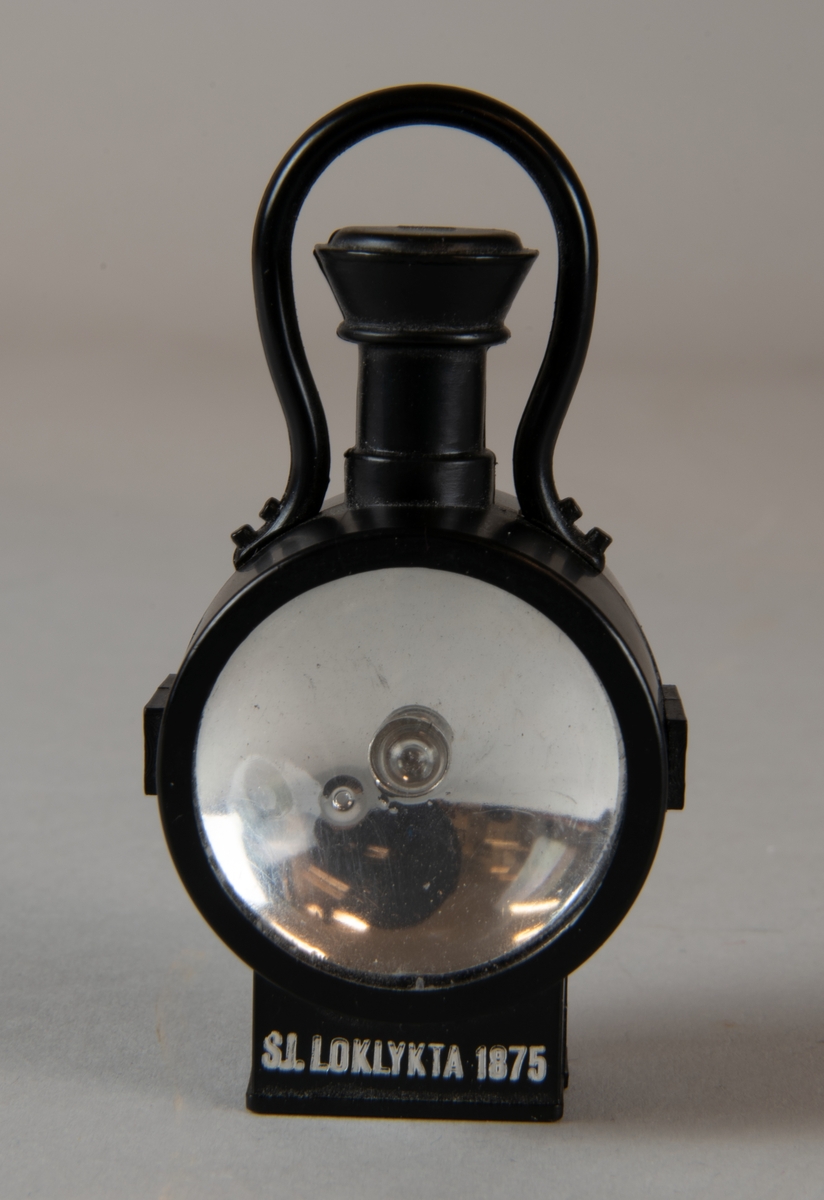 Loklykta i miniatyr av plast med texten "SJ. LOKLYKTA 1875" på framsidan. Loklyktans hölje är svart. I mitten sitter en lampa. Lyktan är batteridriven .