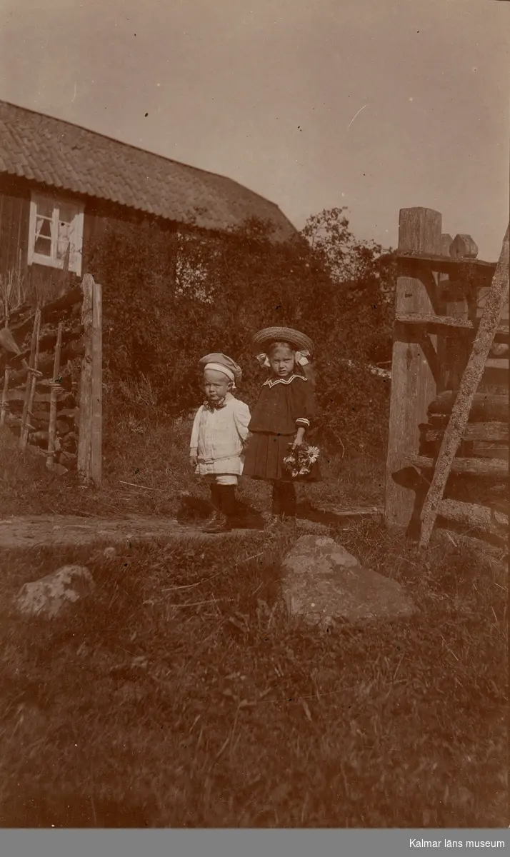Fotografi av två barn utomhus. En pojke Curt Nyström Stoopendaal till vänster? och en flicka till höger. Flickan är klädd i hatt, jacka, kjol, strumpor och skor. Barnen står mitt i en öppning i ett staket. I bakgrunden syns ett hus. I gräset till höger syns stora stenar.