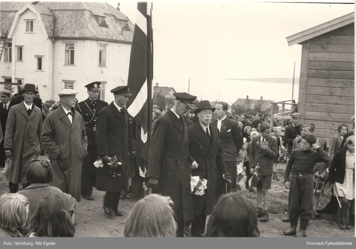Kong Haakon VII på besøk i Vadsø i 1946. Kong Haakon går først i et tog. Til høyre side går fylkesmann Hans Julius Gabrielsen. Mange mennesker har kommet til stede for å se på kongen. Vadsø sykehus (kysthospitalet) i bakgrunnen. Se også bildene 264-267.