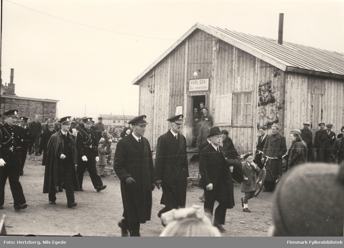 Kong Haakon VII på besøk i Vadsø i 1946. Kong Haakon i midten, høyre siden går fylkesmann Hans Julius Gabrielsen. Mange mennesker står langs veien å ser på. Her går de forbi butikken til Karl Saxi "autorisert installatør". Se også bildene 264-267.