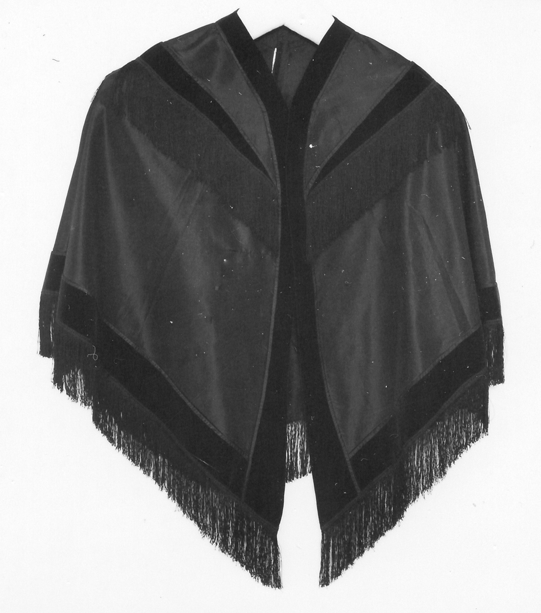 Cape, svart sidensatin. Två tygstycken hopsydda mitt bak. Dekorerad med svarta breda sammetsband och svarta långa fransar. Från omkring 1900.