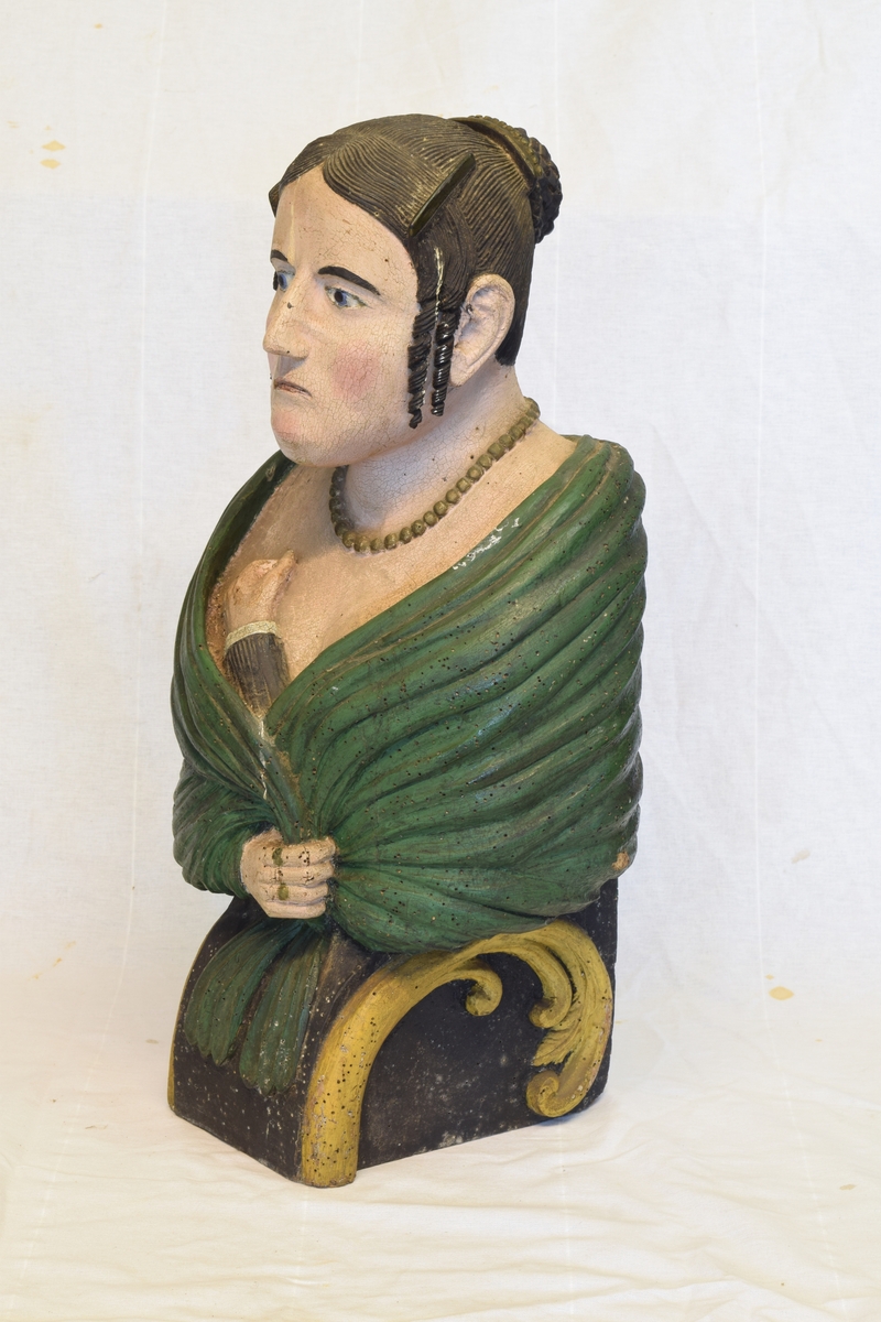Portrettbyste av kvinnehode. Skulle brukes som gallionsfigurer men kan og ha vært brukt som dekorasjon i et hus. Sortmalt hår med hengekrøller ved ørene og grønt sjal.