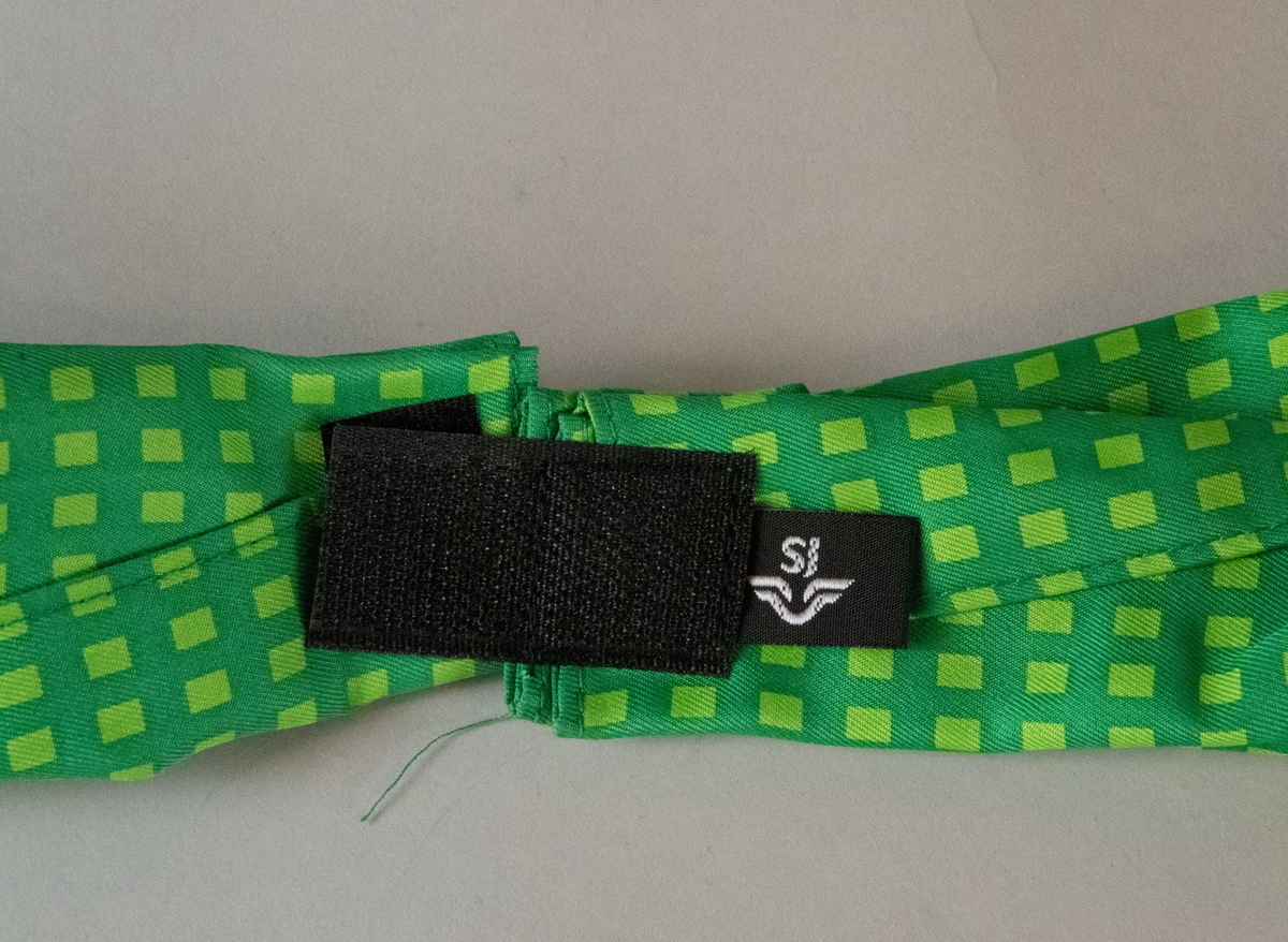 Rektangulär långsmal scarf av grönt tyg, mönstrat med rektangulära små rutor i en ljusare nyans av samma färg. Scarfen är delat i två i nacken, med påsydda kardborrband i ändarna för att sättas ihop. På baksidan fastsytt under ett av kardborrbanden sitter en svart etikett med SJ:s logotyp i vitt, samt tvättråd.
Scarfen är helt oanvänd.