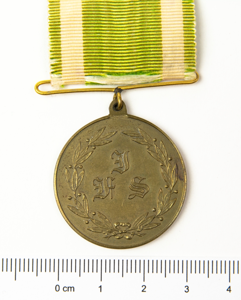 Medalj med band i vitt och grönt. På medaljens ena sida finns en lagerkrans med I F S i mitten. På frånsidan finns en lagerkrans.