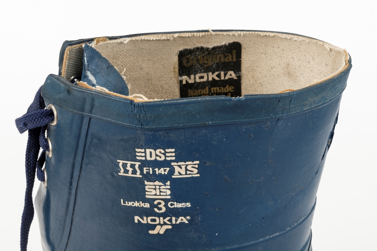Vernestøvler, Nokia i størrelse 43, for skogsarbeid, innkjøpt i 1984.  Alle utvendige deler er utført i gummi, som tilstynelatende er limt eller smeltet sammen, i flere lag der hvor påkjenningen på støvlene var antatt størst, det vil så tå- og hælpartiene og fronten ved vristen og leggen.  I tillegg er ankelkulene beskyttet med sirkelrunde, svarte gummiknapper med stjernemønster.  Den nedre delen av støvlene (fotpartiet) er utført i oransje gummi, den øvre (leggpartiet) i mørkeblå gummi.  Bak på støvleskaftene er det spalter med tre maljepar og lisser som kan strammes slik at åpningen kan justeres tett, slik at skogsarbeideren slipper å få snø i støvlene.  Sålene har cirka fem millimeter høy mønstring, som skal redusere faren for at brukeren sklir.  Sålemønsteret har også hull for pigger, som ikke er innsatt.  Viktig for vernefunksjonen er skjulte stålhetter som er støpt inn i tåpartiet.  Disse komponentene gav beskyttelse både mot uhell med motorsagkjedet og mot eventuelle klemmeskader.  Støvlene har innleggssåler med lær på oversida og et tovet ullmateriale på undersida.  Det innvendige tekstilforet er noe slitt i hælsonene.
