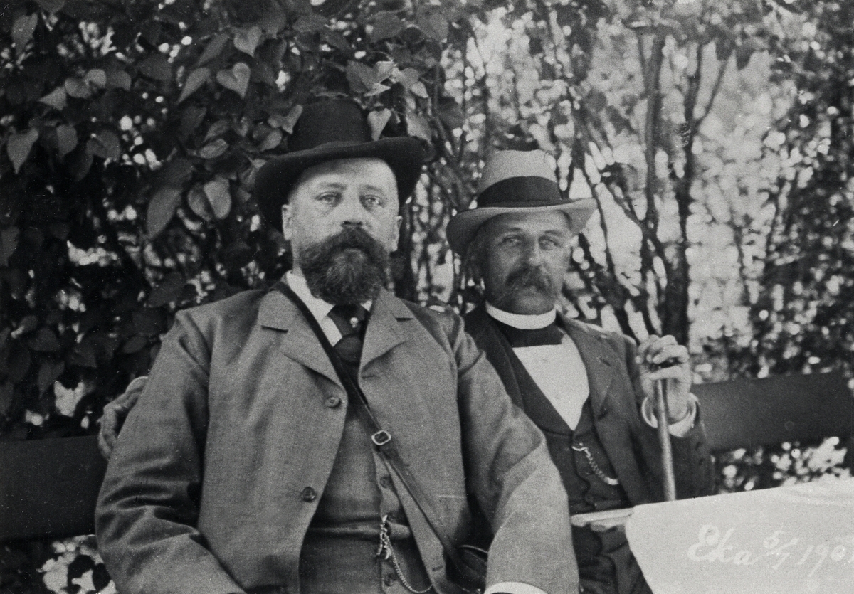 Två herrar i hatt och kostym sitter i en berså. I högra nedre hörnet står: "Eka, 5/7 1901".
Fältläkare m.m. John Nordlund, Stockholm och och hans svåger, kapten/gymnastiklärare Olof Bratt, Växjö.