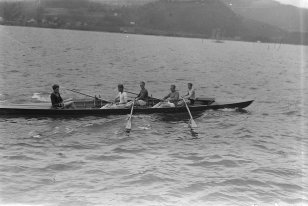 Roing, fire roere og en styrmann i kapproingsbåt på Mjøsa ved Lillehammer, Vingnes i bakgrunnen.