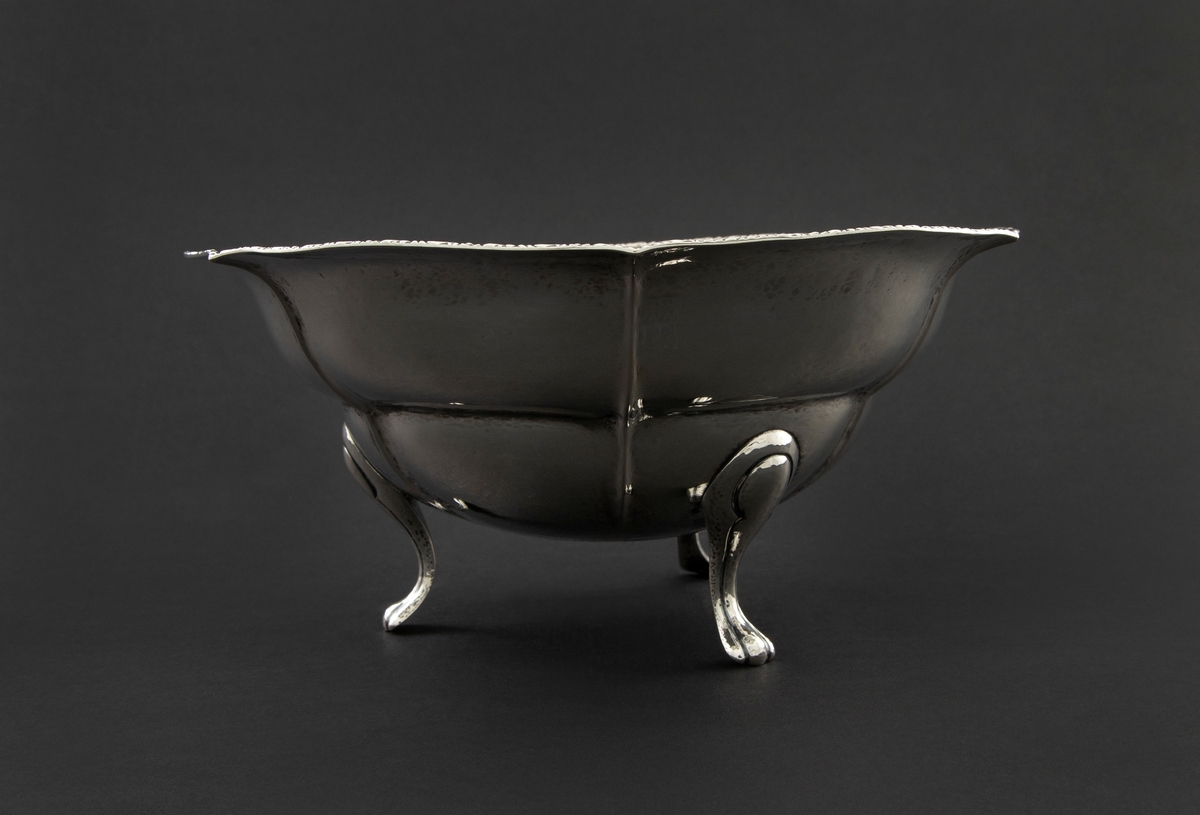Sølvskål med rund, dyp form, bølgende kant og rette knekk. Den står på tre svungne bein. Kanten er dekorert med bonderomantisk ornamentikk.