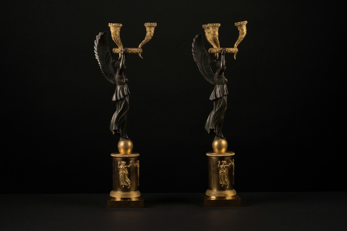 Ett par brännförgyllda bronsljusstakar, kandelabrar, med tre ljushållare vardera.
En kolonnformad bas med ett klot på och en fyrkantig bottenplatta, av brännförgylld brons, bär upp en bevingad kvinna av svärtad brons. I de uppsträckta händerna sitter en krans med tre ymnighetshorn som utgör ljushållarna. Kransen och hållarna är även dessa av brännförgylld brons och dekorerade med akantusblad och vindruvsklasar. 
Runt den runda bärande basen finns reliefer som föreställer tre dansande kvinnor, de tre gracerna, som håller i blomstergirlanger/festonger.