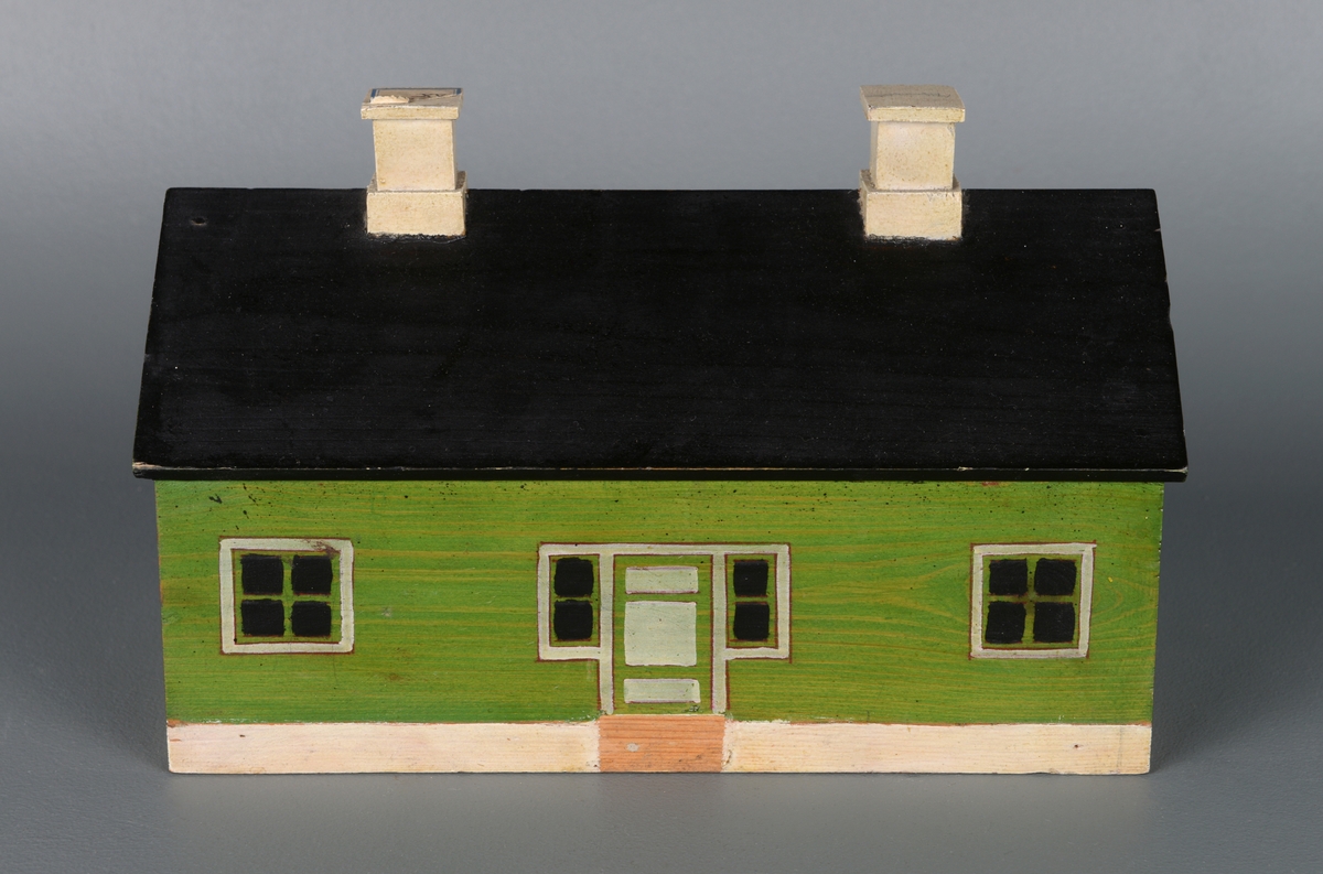 En liten modell av et hus laget av re og montert på en treplate. Huset er rektangulært med skrått tak. Det har to piper, tre dører og 12 vinduer og en liten trapp. Huset er malt grønt, med hvite karmer og hvite piper.