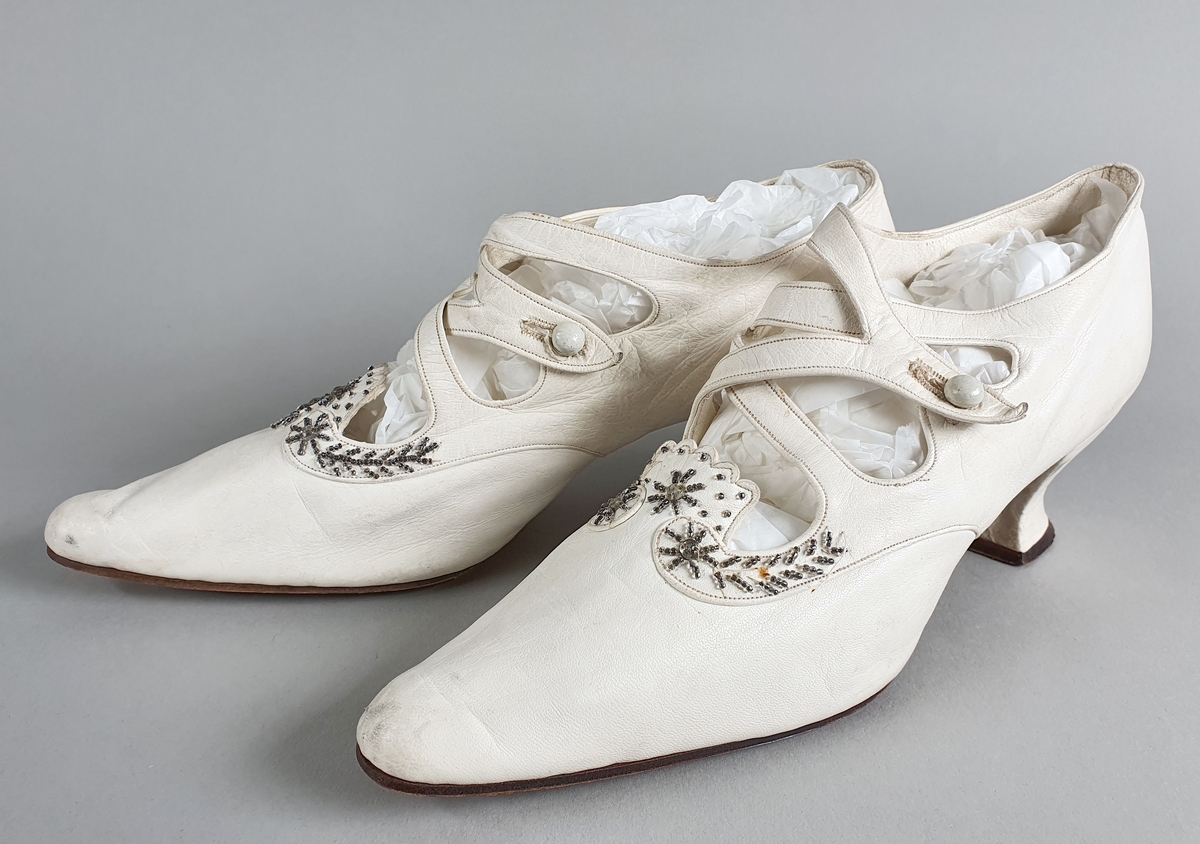 Hvite brudesko av mykt skinn med reimer og dekor av sølvperler over vristen, og med knapp på hver side. Skoene har middels høye hæler som skråner inn under sålen.