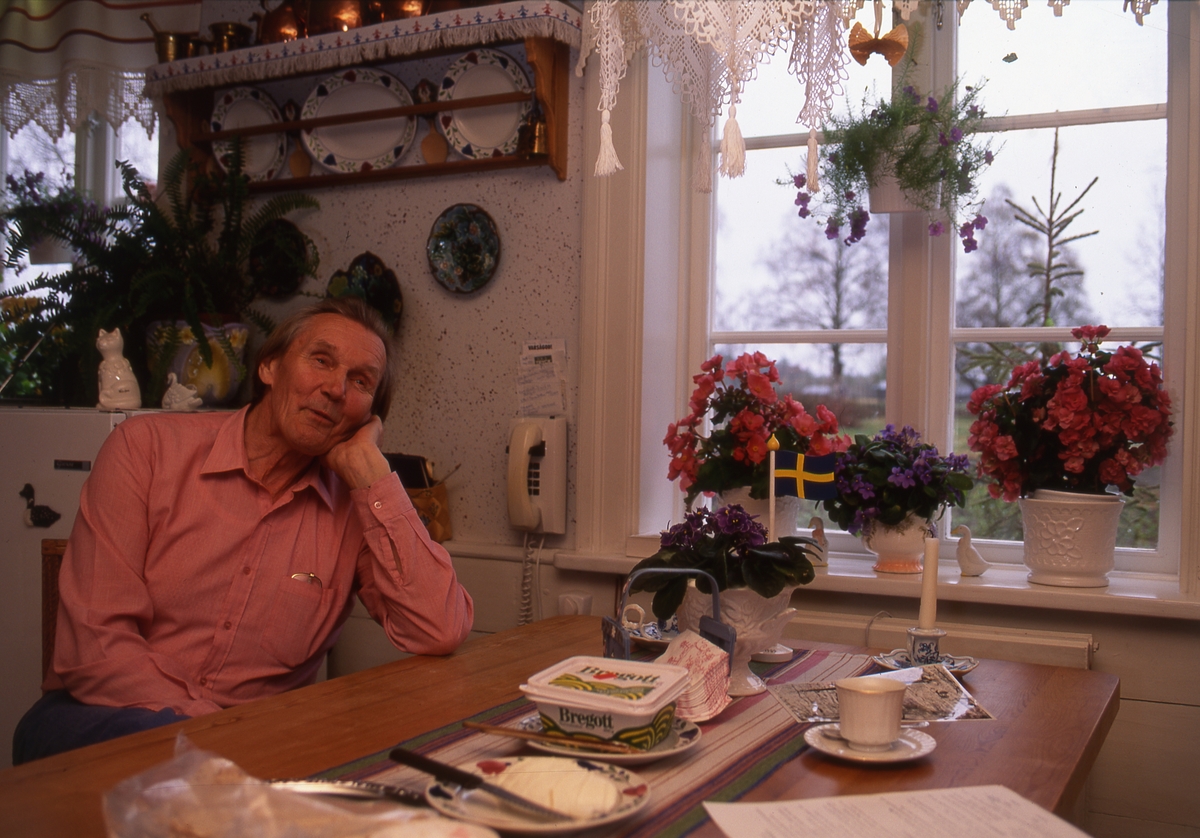 Hilding Mickelsson sitter vid matbordet hemma i köket i Sunnanåker. Han lutar huvudet i ena handen och ser ut att prata med någon. På bordet är smörgås och kaffekopp framdukad till en person.