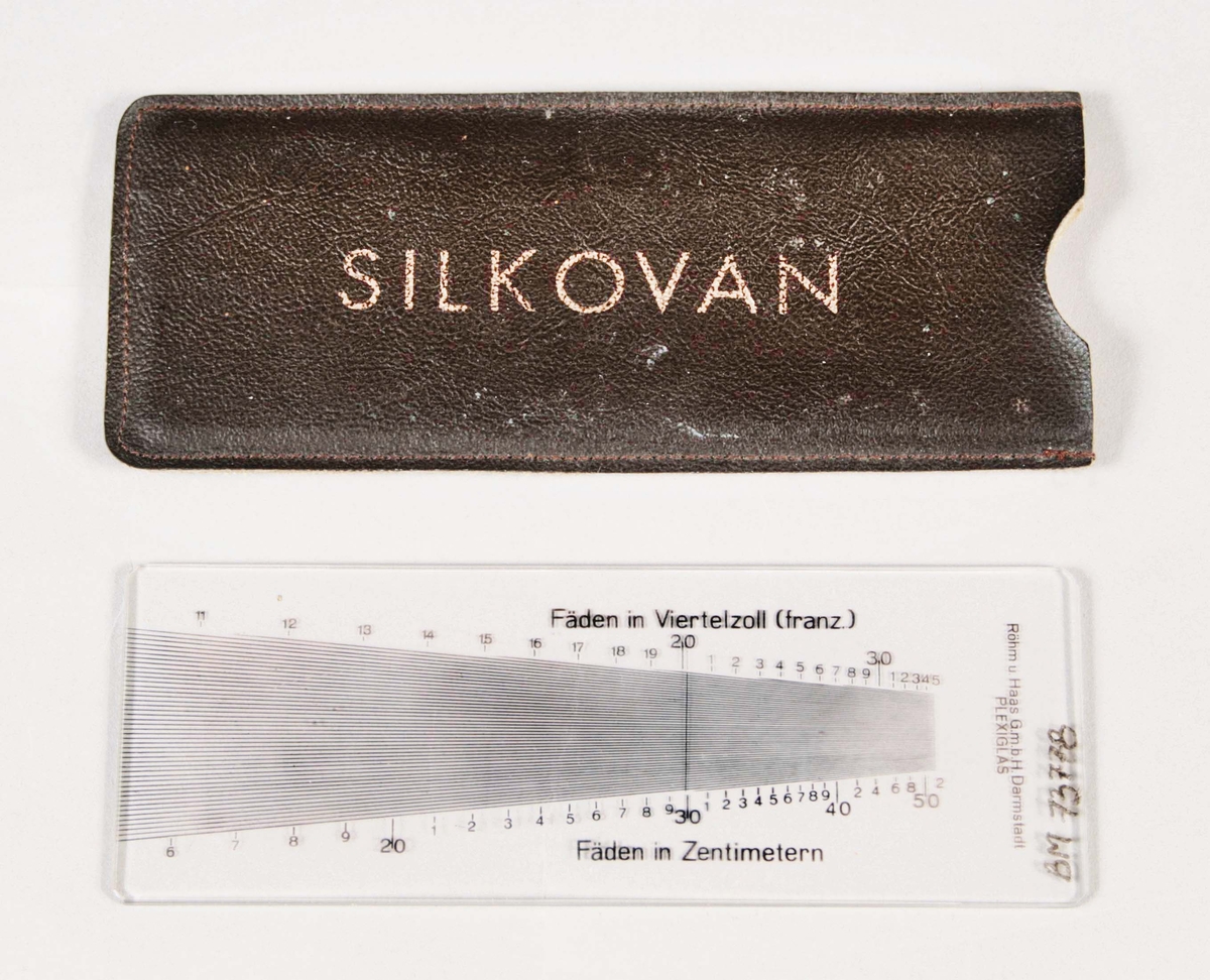 Måttinstrument. Plexiglasskiva, med svarta skalor, för att se "Fäden in Zentimetern" och "Fäden in Viertelzoll (frannz." I svart galonfodral med guldtryckt text, märkt "SILKOVAN".