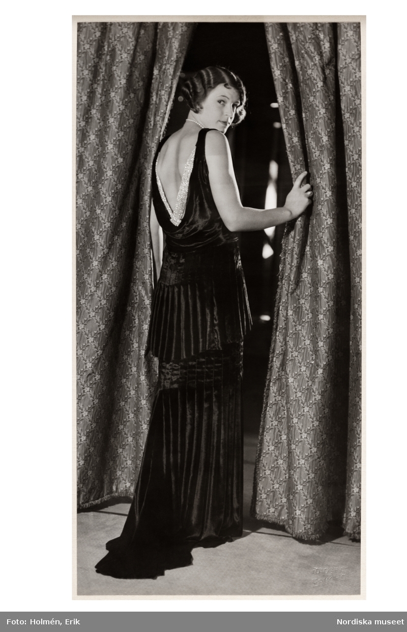 Mannekäng visar mode. Festklänning med ryggen i fokus, ärmlös klänning med urringad, draperad rygg dekorerad med glittrande tyg. Hellång med veckad skört och kjol med rak silhuett, litet släp. NK:s Franska damskrädder. hösten 1930i, från Jeanne Lanvin.