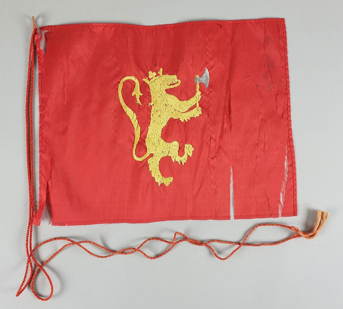 Rødt silkeflagg med brodert løve i gult. Handfallet kant på tre sider, jarekant på en side. I overkant en tvunnen snor