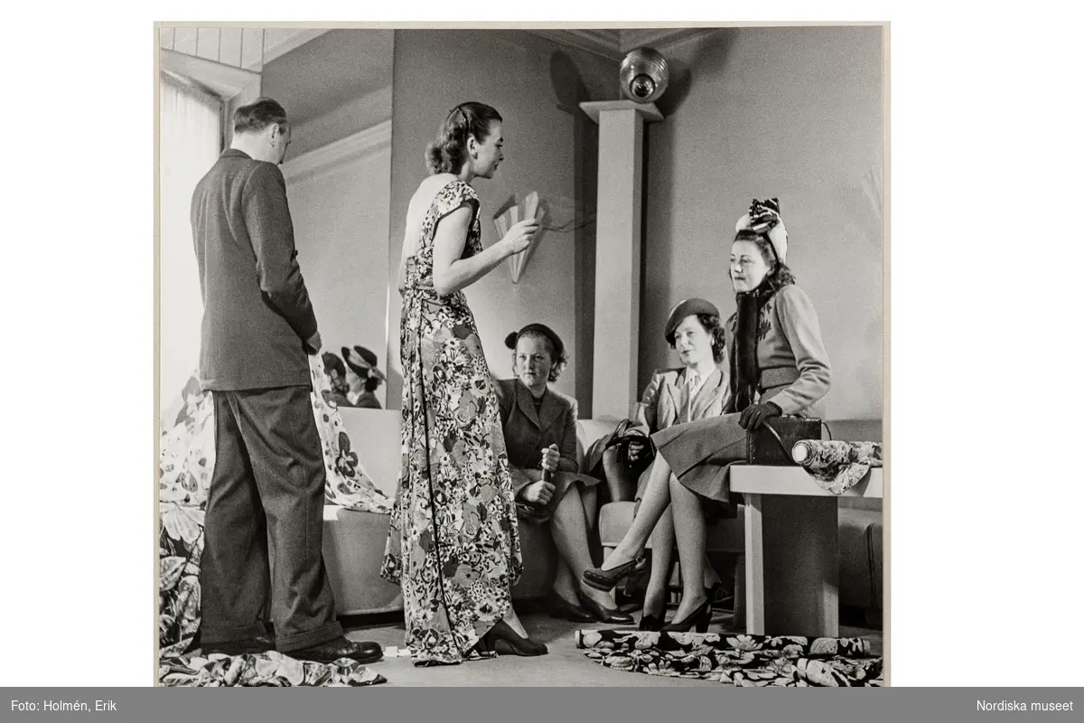 Mannekäng visar mönstrad klänning ur sommarmodet för tre kvinnliga kunder sittande i soffa i salongen på NK:s Franska damskrädderi, Bakom henne står Pelle Lundgren. På golvet ligger tyger.