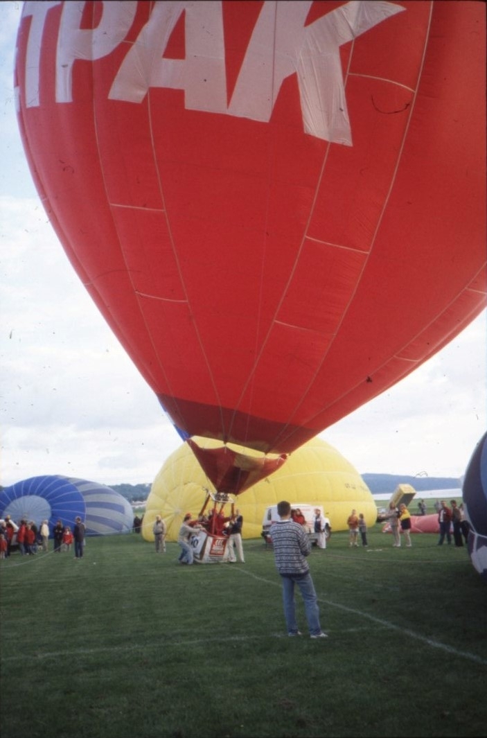 En röd luftballong påväg att lyfta från marken. I bakgrunden ses två till hälften fyllda luftballonger. Två personer befinner sig i korgen medan minst två personer står bredvid och håller i korgen.