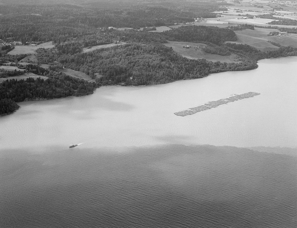 Tømmerslep over innsjøen Øyeren sommeren 1985.  Tømmer fra Østerdalen og Solør, Gudbrandsdalen, Mjøstraktene og bygdene på Øvre Romerike ble fløtet løst ned til Fetsund lenser i nordenden av denne innsjøen.  Her ble virket fra gammelt av sortert og «soppet» eller «moset» (buntet), og det som skulle til Lillestrøm, Strømmen og Oslo ble skilt fra det som skulle videre nedover vassdraget.  Da dette fotografiet ble tatt var det bare storbedriften Borregaard fabrikker i Sarpsborg som fortsatt fløtet, så noe særlig sortering var det ikke snakk om lenger.  Tømmeret ble moset i høvelige bunter med hver sin «grime» (vaierbindsel).  Inntil 200 av disse mosene ble bundet sammen i ei «vending» som ble buksert forsiktig ned gjennom en meandrerende djupål i det grunne deltapartiet lengst nord i innsjøen.  Når ei slik vending kom fram til Sofiedal ved Engelsvika på innsjøens vestside, like ved grensa mellom Rælingen og Enebakk kommuner, ble den ankret opp og slepebåten Mørkfos gikk tilbake til Fetsund for å hente nok ei vending av samme type.  Når også dette var framme ved Sofiedal ble de to slepene koplet sammen, slik at Mørkfos kunne trekke begge to sørover med ei lang trosse.  I noen tilfeller ble det også hektet på bommer med løstømmer fra «etterrensken» langs sjøen.  Slepet kom gjerne fram til Sleppetangen, ved nedre Glommas utløp fra Øyeren, seint på kvelden.  Da ble slepet oppankret mot land, slik at lokale fløtere påfølgende dag kunne løsne vaierbindslene og la tømmeret flyte løst nedover mot tømmerrennene forbi kraftverksdammene nedover i vassdraget.  Mørkfos gikk nordover mot Fetsund igjen om natta, slik at båten kunne fortsette slepearabeidet derfra neste morgen.  Dette flyfotografiet er tatt etter at to slep var koplet sammen til ett, altså nedenfor Sofiedal, idet det slepes langs Øyerens vestbredd.  Opptaket ble gjort i 1985, det siste året det foregikk kommersiell tømmerfløting i dette vassdraget.