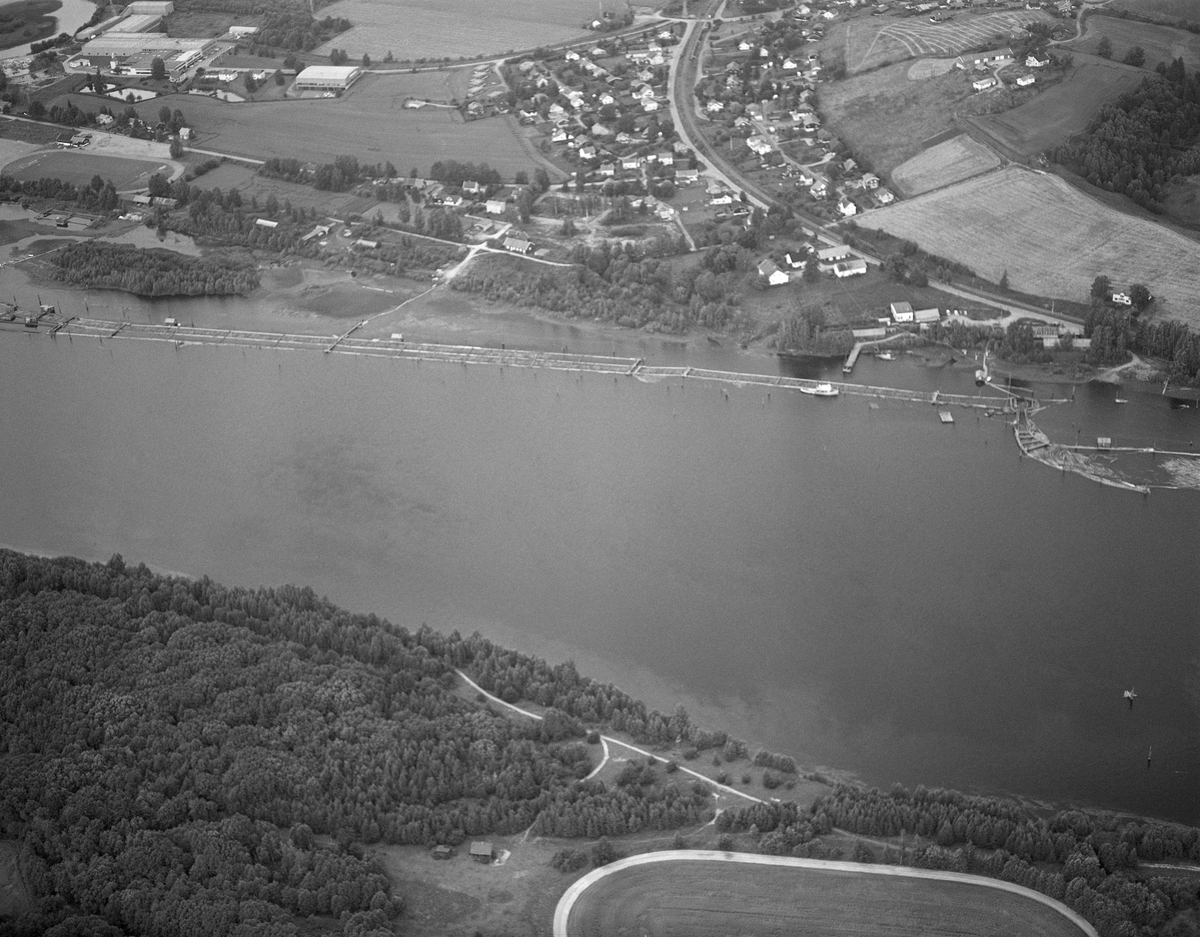 Flyfotografi, tatt over deler av lenseanlegget på Fetsund i Fet kommune, Akershus i 1985. Bildet er tatt fra en posisjon over den østre elvebredden, på tvers av elveløpet, som rant fra høyre mot venstre når det betraktes fra øst. Tømmeret ble sluppet porsjonsvis ned mot Fetsund fra attholdslensa ved Bingen, sju-åtte kilometer lengre nord. Da dette fotografiet ble tatt, i Glomma-fløtingas sluttfase, var det bare Lundlensa ved den vestre elvebredden som fortsatt var i drift på Fetsund. Tømmeret ble ledet dit via ei traktformet lense som lå under jernbane- og vegbruene. Nederst i denne inntakssona var det en knekk på det flytende stengselet (til høyre på dette bildet). Der ble løstømmeret løftet over ei bru med roterende jernvalser og kastet paralelt ned i «tømmerrenna», en rettlinjet kanal omgitt av flåteganger. Her fløt stokkene langsomt nedover mot bunteanlegget - «soppemaskinene» - i den nedre enden av lenseanlegget, helt til venstre på dette fotografiet. På den vestre elvebredden lå Glomma fellesfløtingsforenings eiendom Lund, med lensebestyrerbolig, kontorer og en rekke økonomibygninger. I fløtingas avviklingsfase var det her virksomheten hadde sin hovedadministrasjon - tidligere hadde den adresse i Osloi, 1985 var det siste året det ble fløtet tømmer i Glomma, og dette året var det bare hovedvassdraget fra Rena i Åmot og sørover som hadde fløting. Fløtingskvantumet hadde et volum på 126 014 kubikkmeter, som var et svært beskjedent tall sammenliknet med hva som var vanlig i dette vassdraget en generasjon eller to tidligere.
