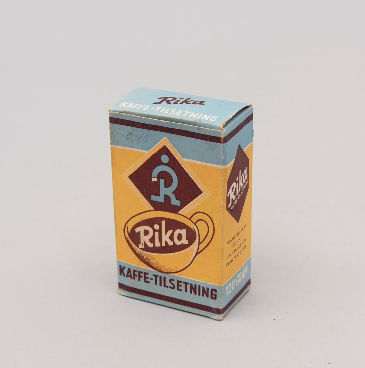 "Rika. Kaffe-tilsetning". Pappeske med innhold i matpapir. Påskrevet."0,40" (pris) med gråblyant. Produsert av A/S P.G. Rieber & Søn, Bergen. (motiv i gult, blått og brunt)