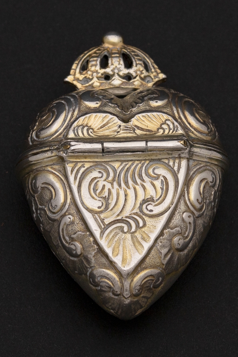Hjerteformet luktevannshus i sølv med gjennombrutt krone på lokket. Rokokko-ornamentikk på korpus og hjerteformet felt på begge flate sidene. Inn- og utvendig forgylt. Beholderens øverste kant er bølgeformet.