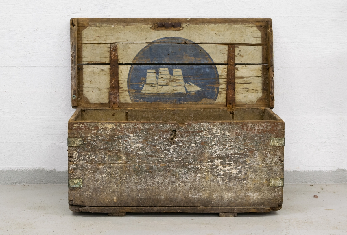 Kiste med seilskute motiv på innsiden av lokket. Falmet og avskallet maling på utside. Håndtak på en av kortsidene.