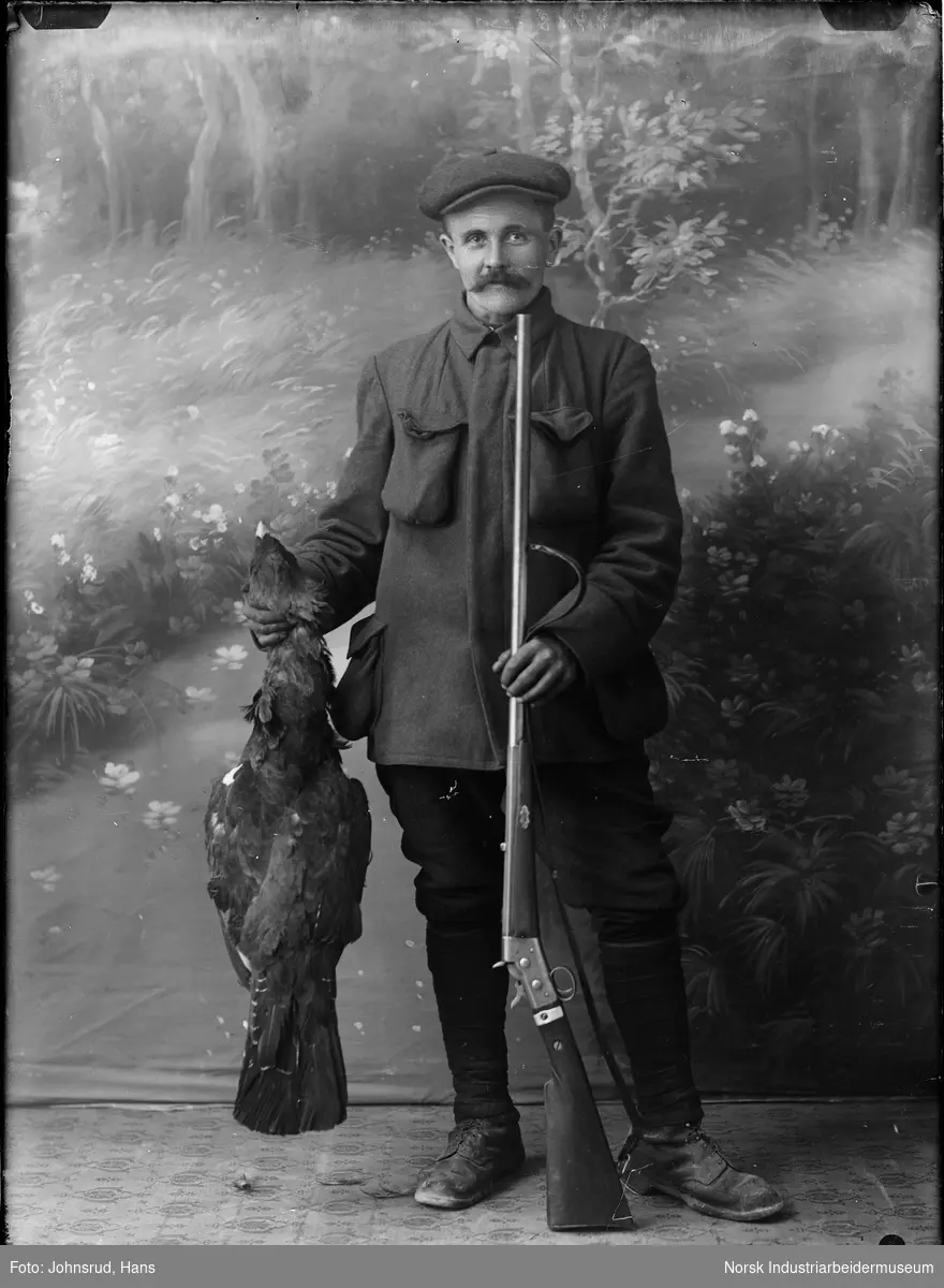 Portrett av stående mann med lue som holder gevær og død fugl.