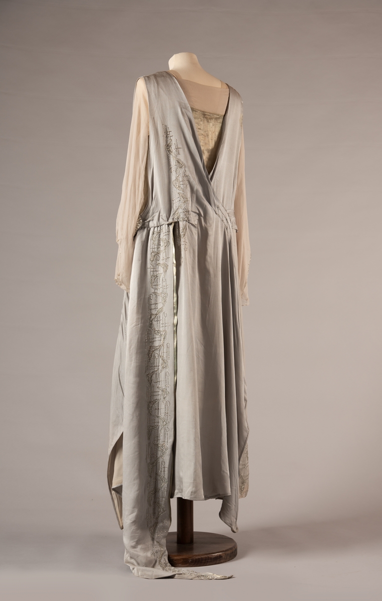 Selskapskjole i grått silkestoff med hvitt fôr i silkestoff. Kjolen er dekorert med glassperler (strass). Kjolen er produsert av Paulsson & Co i Oslo, trolig på 1920-tallet.