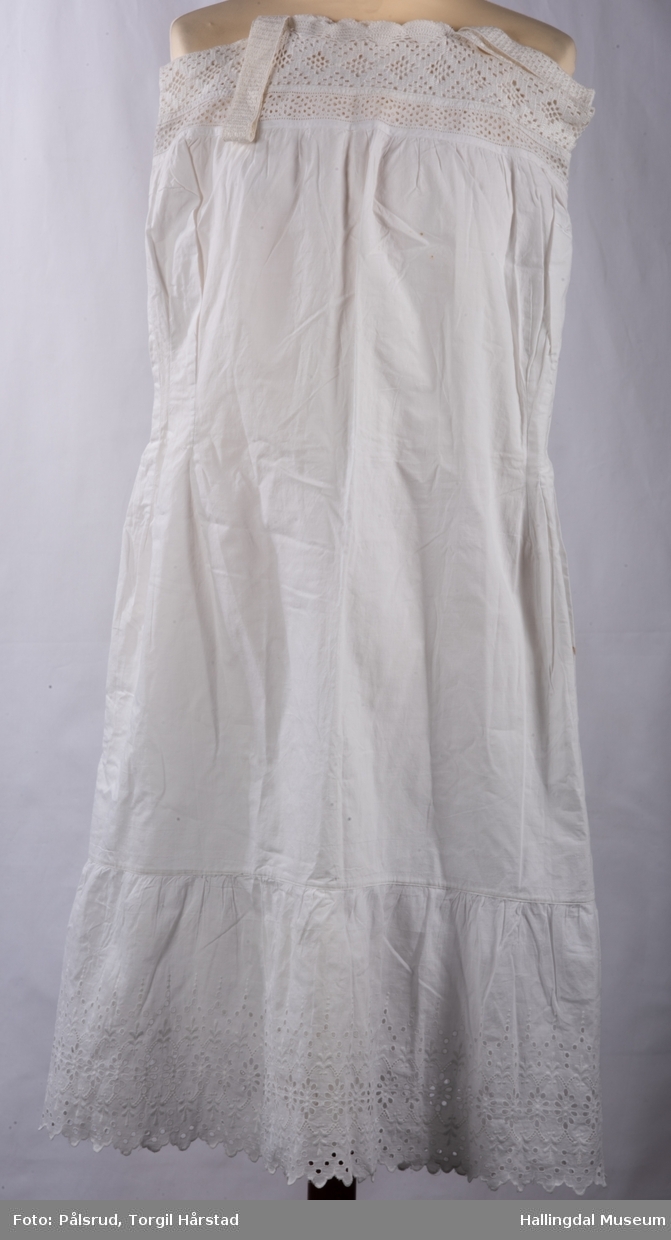 Hvit underkjole i bomull med bred heklet kant øverst på brystet rundt hals og ermer. To heklet stropper. Bred heklet kant nederst på kjolen. Fasongsydd.