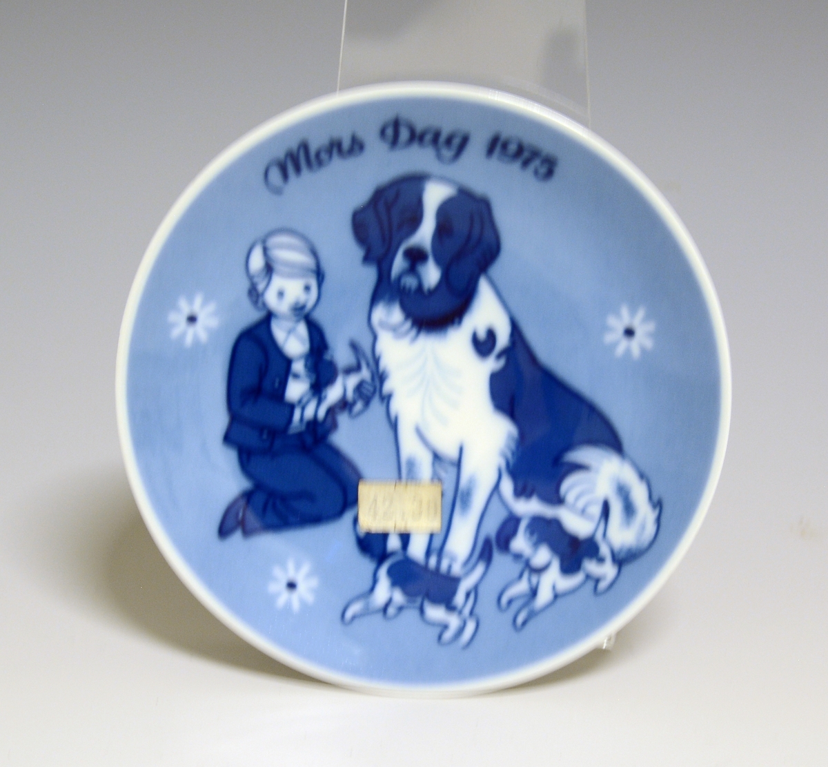 Morsdagsplatte av porselen. Hull til oppheng bak. Hvit glasur. Dekorert i blått med en gutt med hund og valper, tekst Mors Dag 1975.