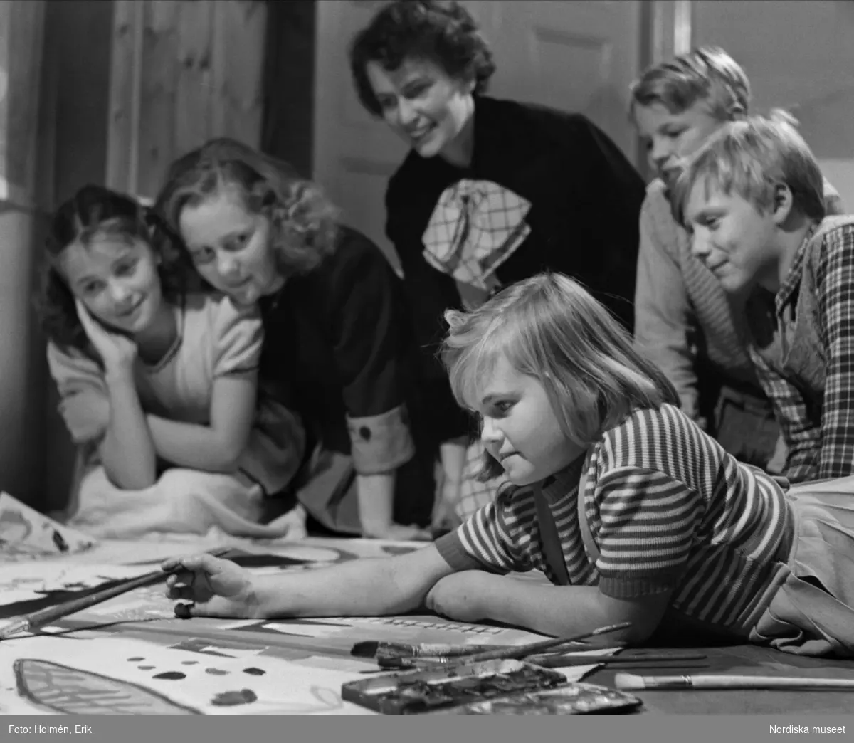 Flicka målar dekor till utställningen "Barnens värld" på Nordiska kompaniet 1949, kvinna och andra barn ser på.