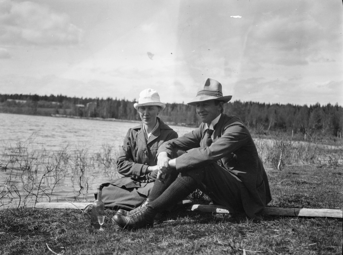 En mann og dame som sitter i gresset ved en innsjø. Flaske og et dramglass foran