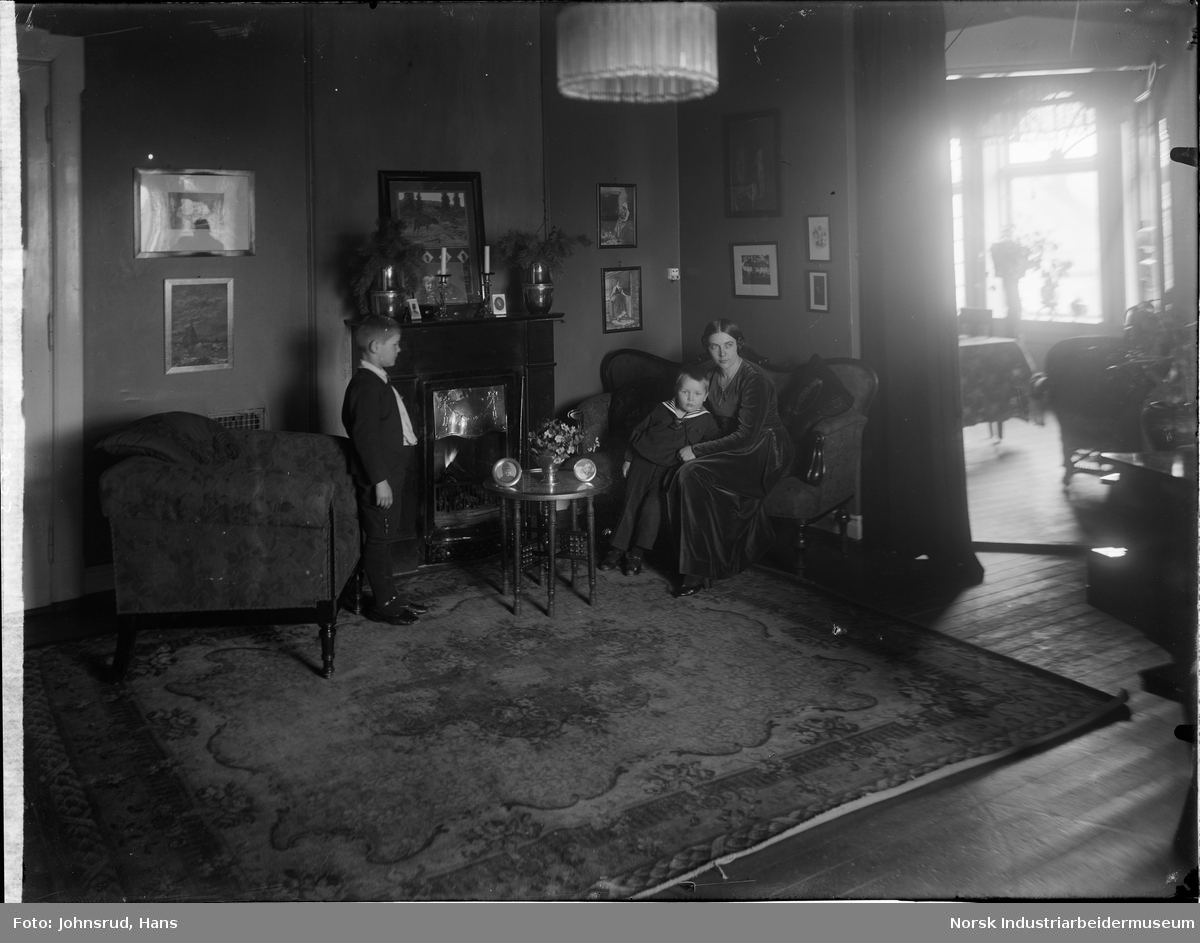 En sittende kvinne og to gutter, en stående og en sittende, i en sittegruppe rundt en peis. Dørgang til et annet rom er synlig på høyresiden av bildet.