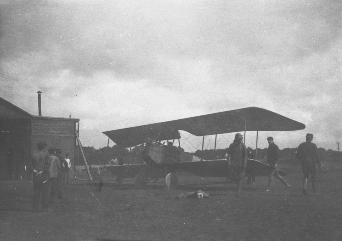 Ett flygplan av typ Thulin på ett flygfält vid hangar. Flygare och personal bredvid flygplanet. Omkring år 1916. Med annorlunda motorinstallation. Flygplan typ Thulin E eller flygplan typ Thulin L.