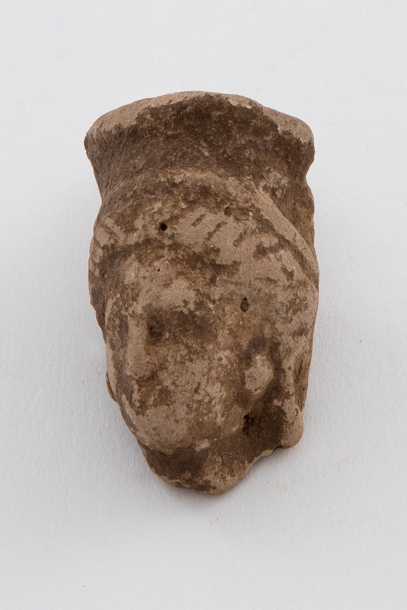 Fragment av figurin i terrakotta. Kvinnehode med hodeplagg med ansiktet i tre fjerdedels profil. Grågult gods.