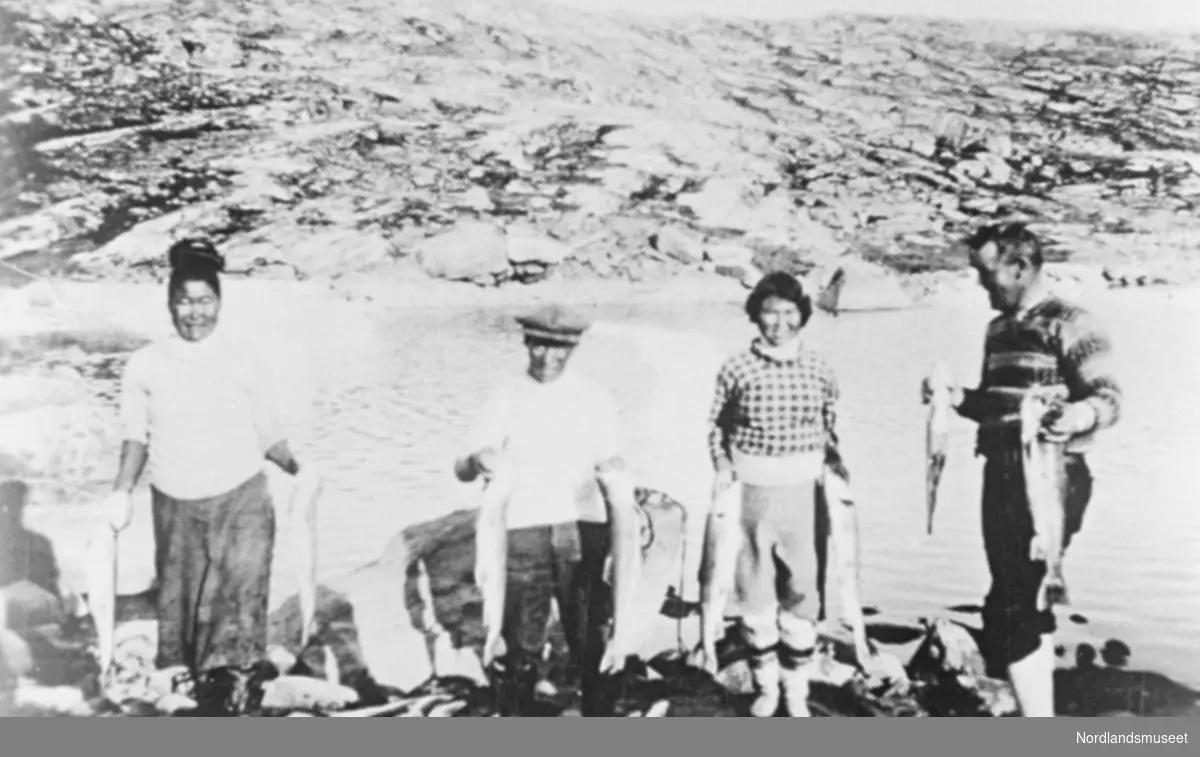"Grønlandslaks" fra Franz Josefs land, Grønland. 4 personer, alle med 1 laks i hver hand. Havet og bergskråning lenger bak.