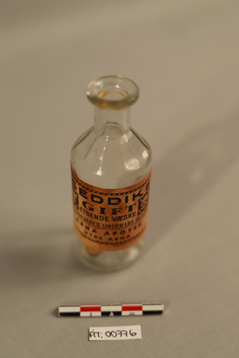 Klar glassflaske med beige, møkkete etikett. Sort skrift på etiketten med to gift logoer (Sort firkant med hodeskalle i).  Rester av korken inni flasken. 