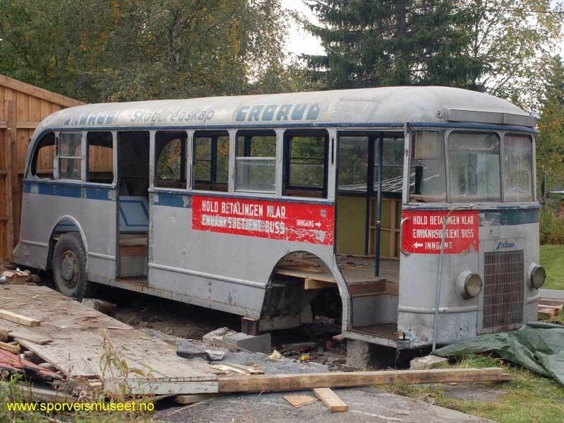 Sølvgrå buss med et mørkt blått bånd langs midten. Bussen mangler dører og noen vinduer, og under og over vinduene er det plassert reklame eller informasjonsskilt. 