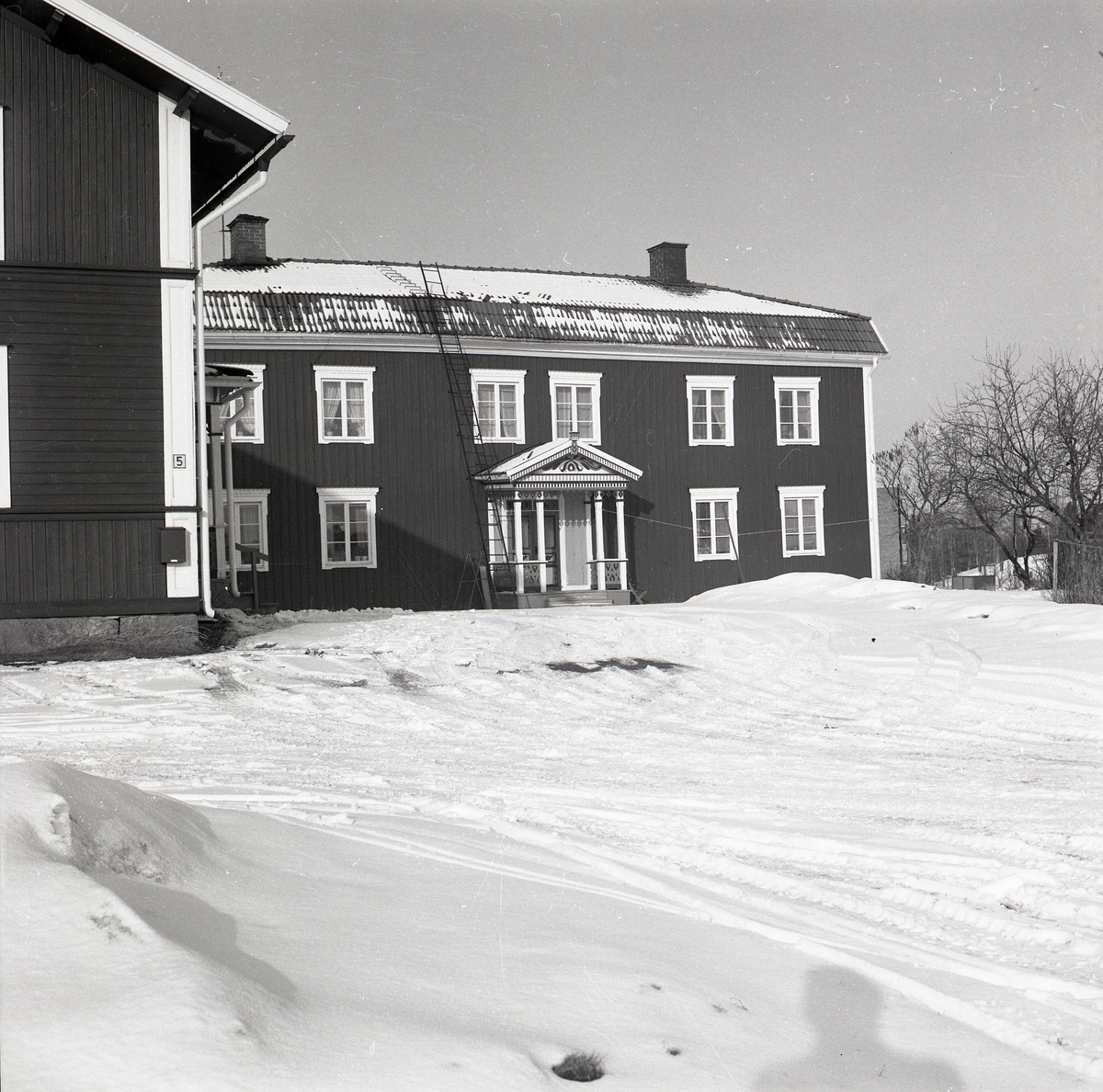En gård med två byggnader i Edsbyn 4 april 1979. Huvudbyggnad med förstukvist och valmat tak. På den snötäckta marken syns skuggan av fotografen.
