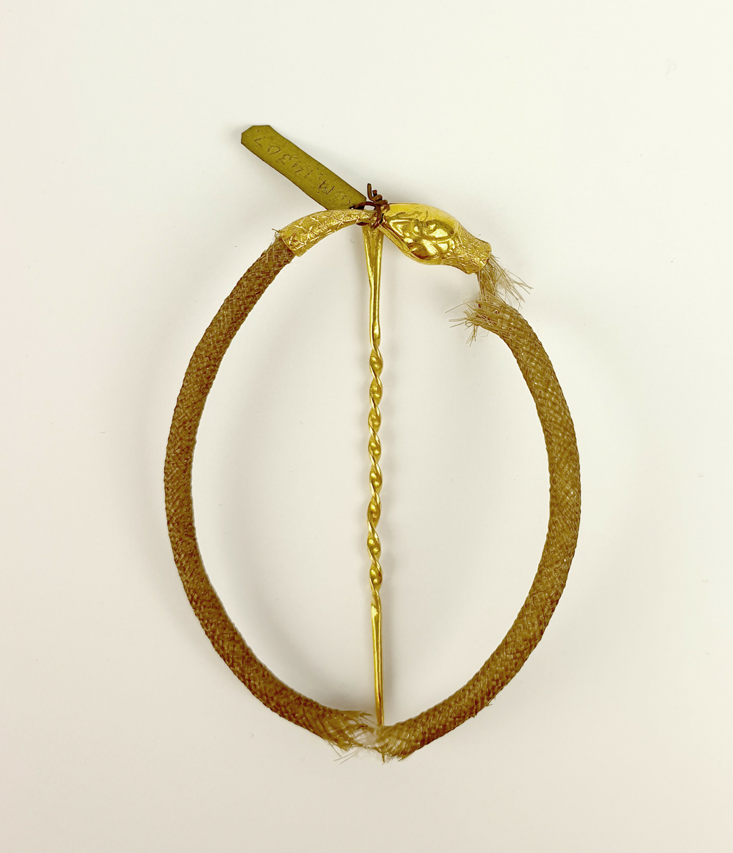 Prydnadsnål, nål av guld. Tillverkad av Arvid Öhrn, Gävle.
Hårarbete: form av en orm. Huvud och stjärt av guld, likaså nålen. Stämplad G 18K m.m.