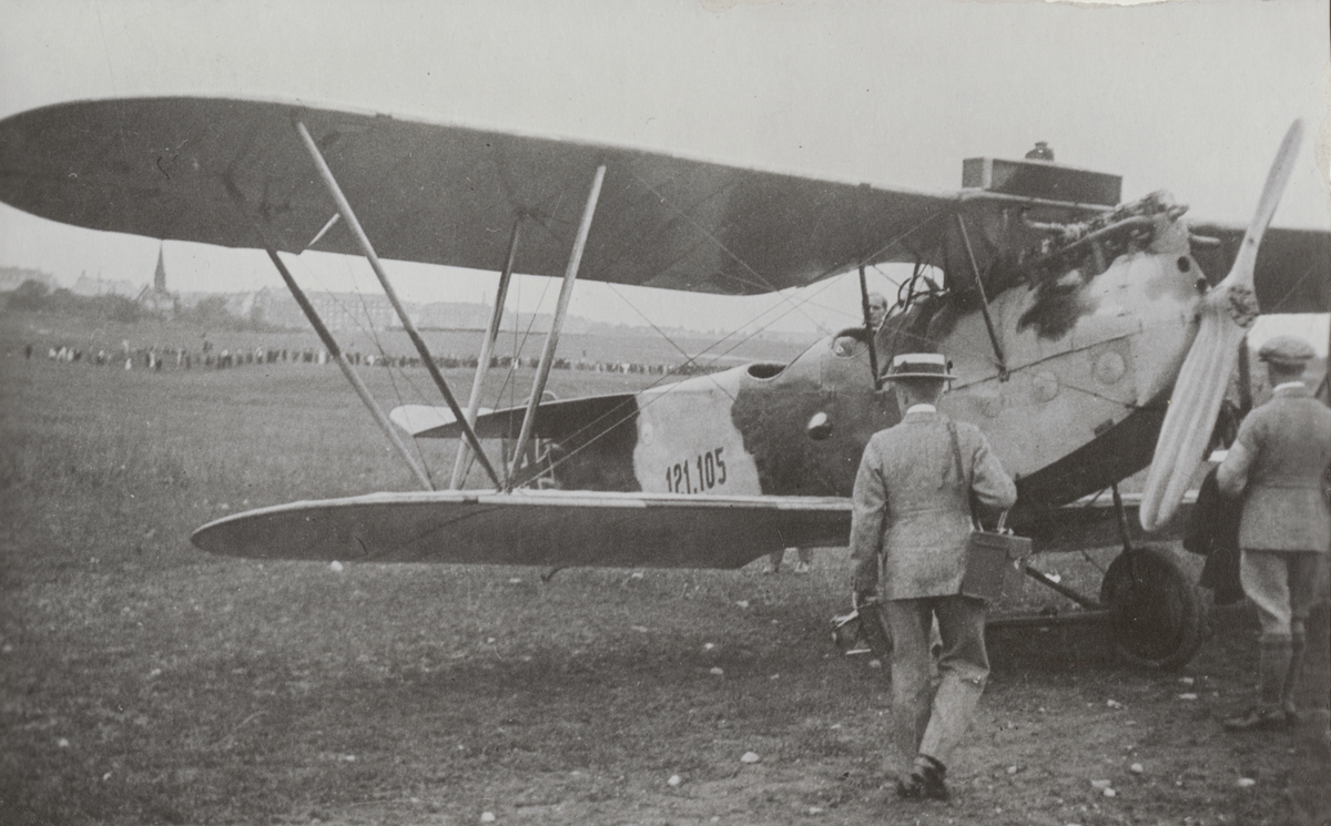 Flygplan Phönix C.1 Dront med tysk märkning 121.105 står på flygfältet på Malmen, juni 1919. Senare "Ur-Dronten" i det svenska försvaret. Ett par män står vid flygplanet. I bakgrunden står en folksamling.