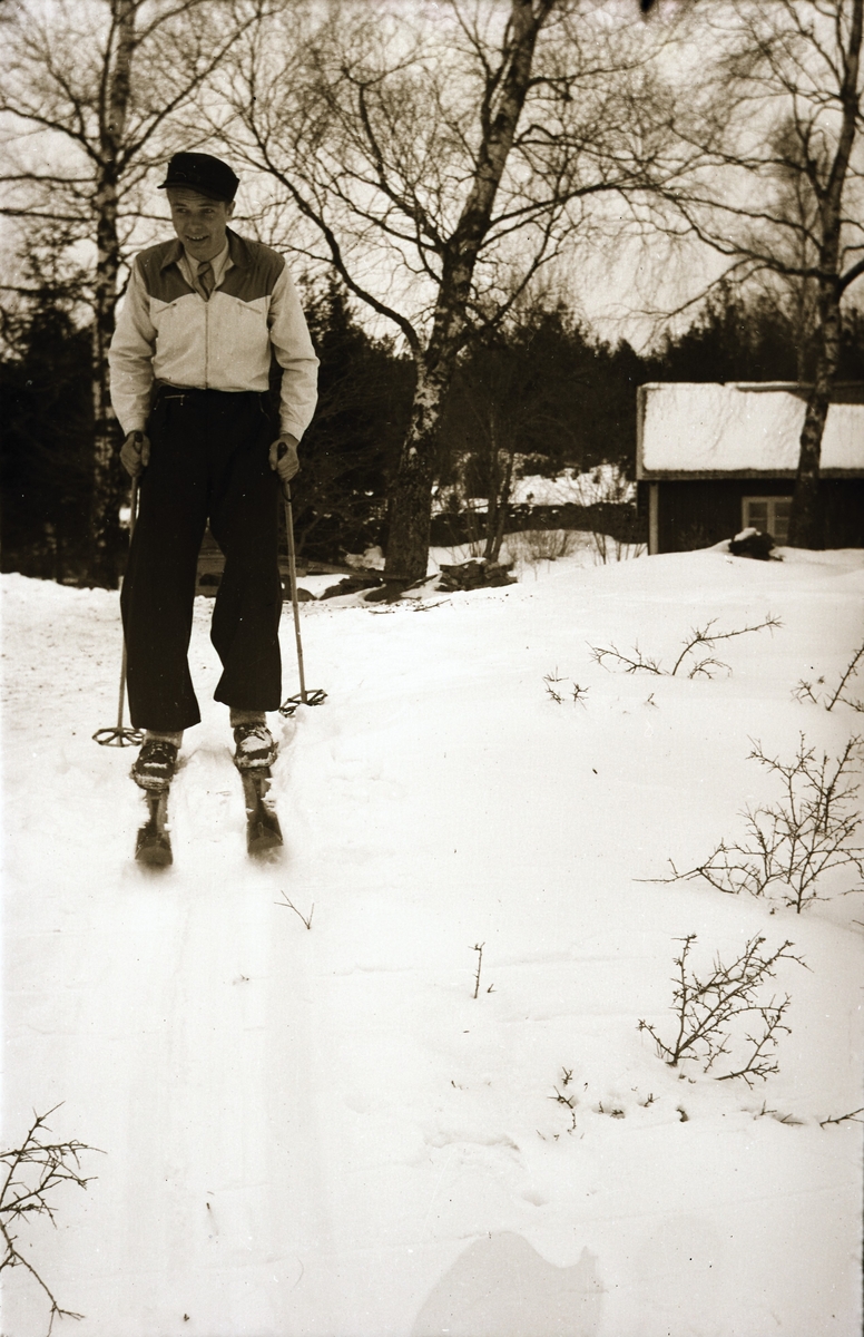 Kjell Börjesson på skidor utför en liten backe. I bakgrunden ligger ett torp.