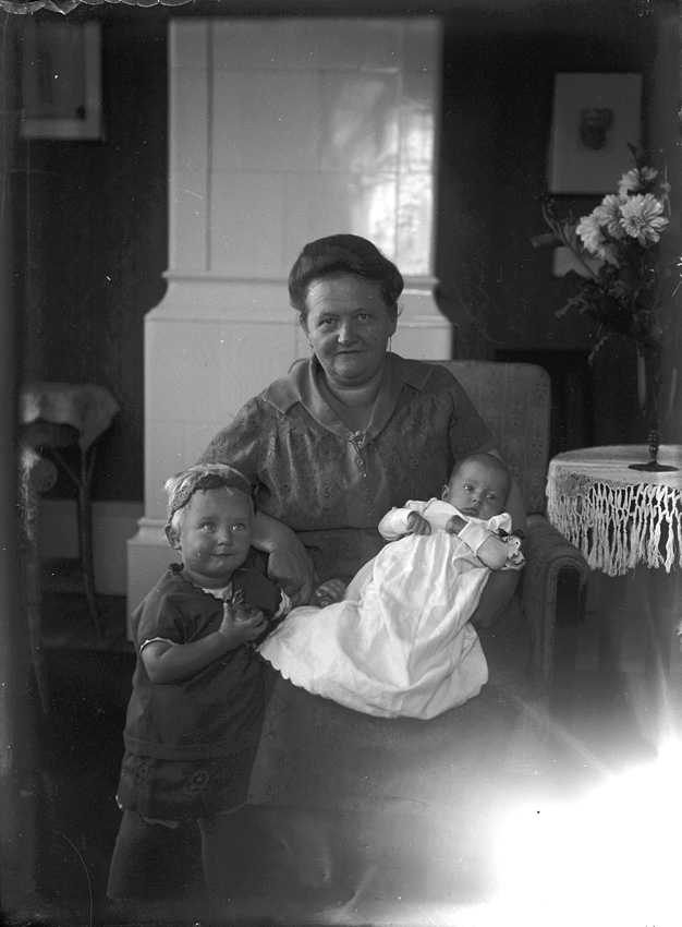 Sittande kvinna med ett barn stående bredvid sig och en baby, klädd i dopklänning, i knät.
Västerås.